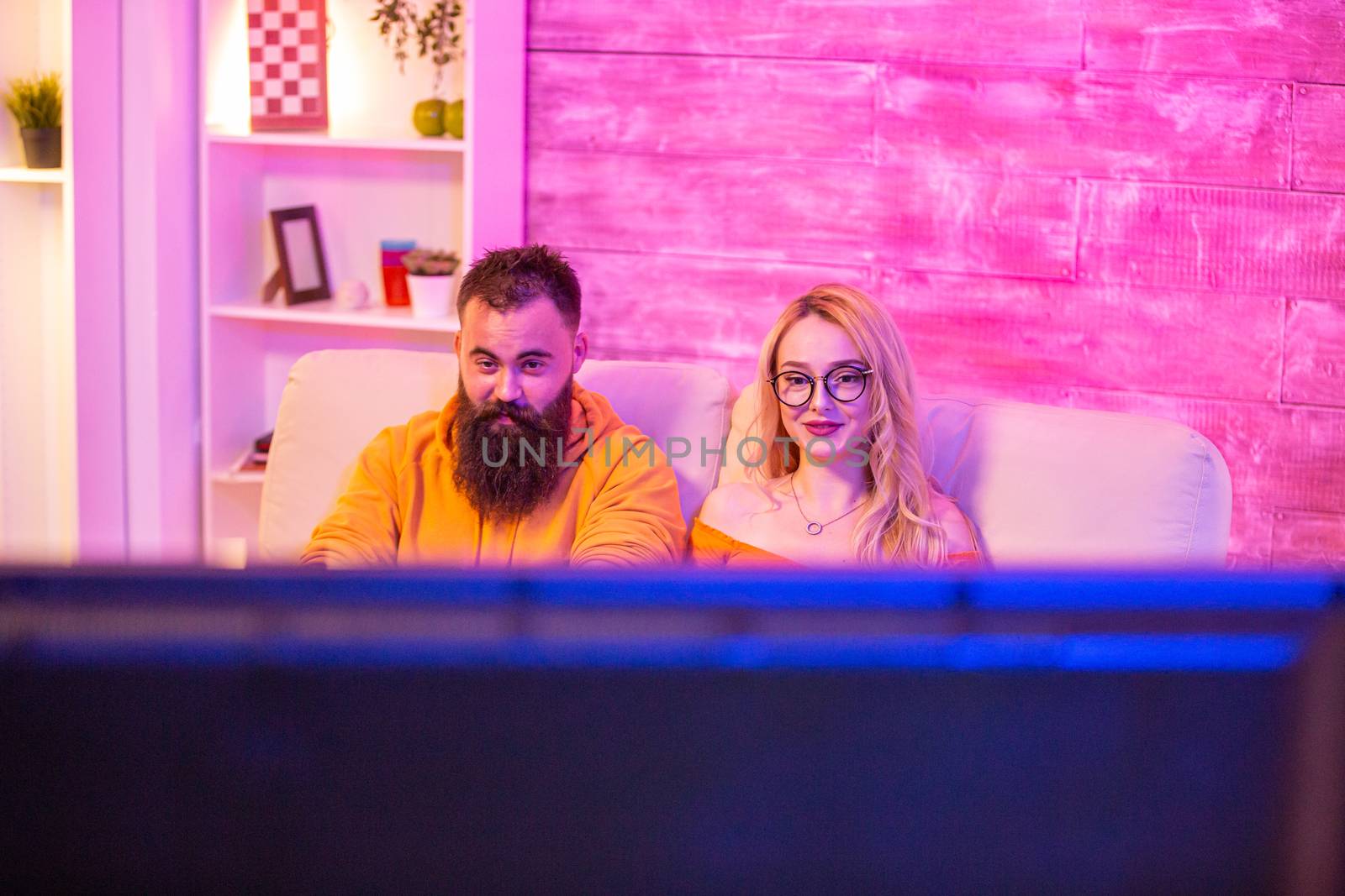 Beautiful blonde girl smiling while playing video games with her boyfriend using wireless controllers on a big screen TV