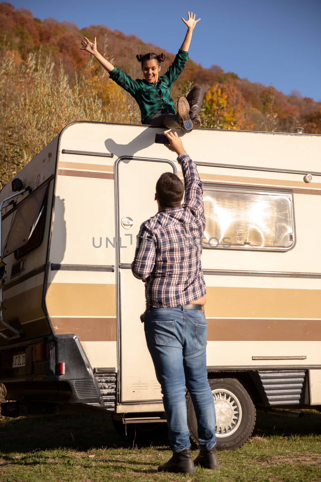 Young man taking a picture of his girlfriend sitting on the roof of a retro camper. Happy time spent together