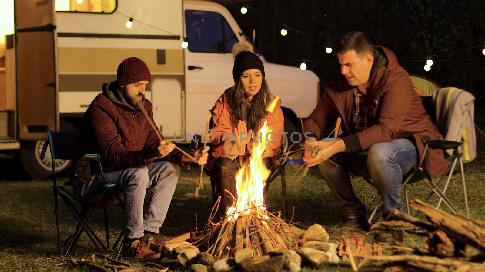 Girl warming up her hands while her friend making the fire stronger. Camp fire. Retro camper. Light bulbs.