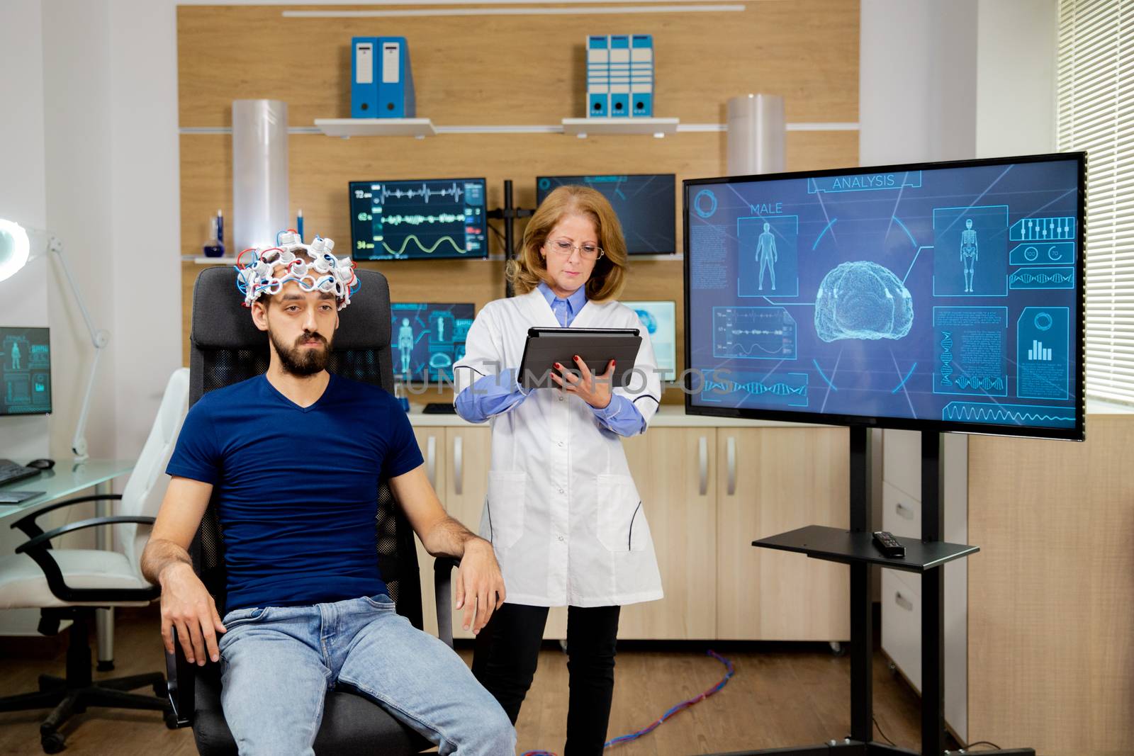 Doctor who follows a male patient during brain scan procedures and makes notes on the tablet. Brain waves scanning helmet