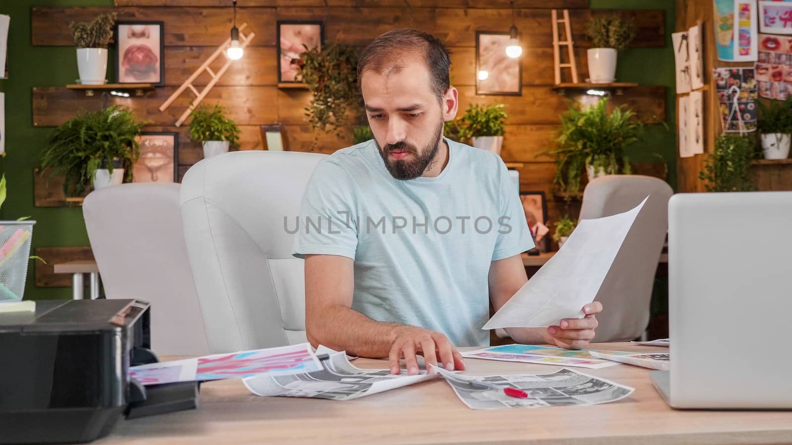 A designer boy sitting at the table and looking at some printed pictures. Creativ office