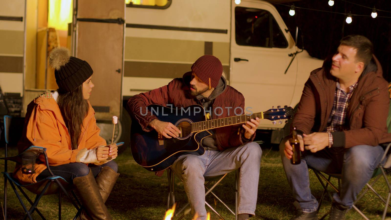 Girl holding marshmallow and one of his her friends is singing on a guitar around camp fire in cold night of autumn. Camping in mountains.