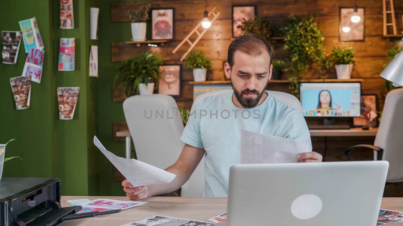 Young designer looking at sheets sitting at his desk with laptop in front. Cozy work space