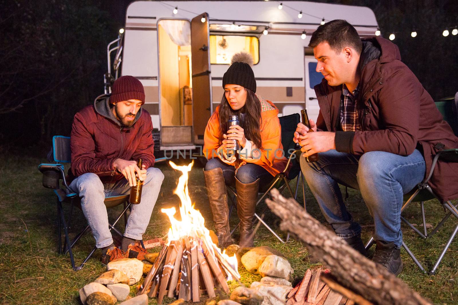 Close friends sitting together on camping chairs around camp fire in the mountains. Retro camper van.