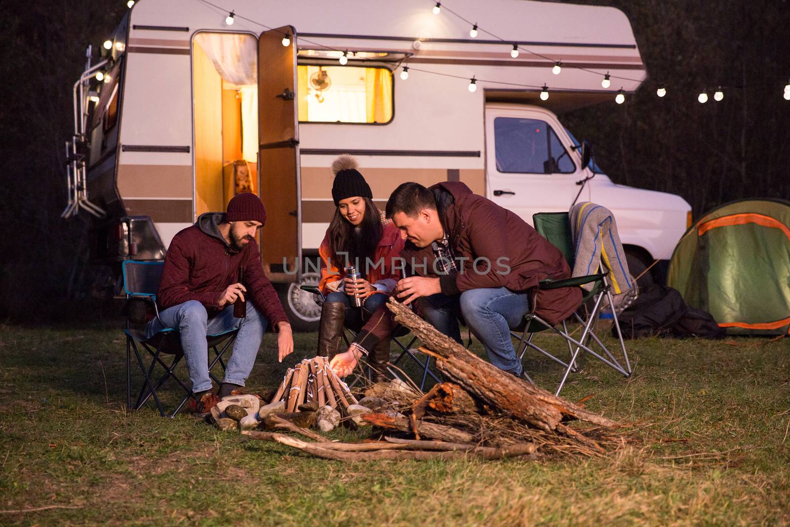 Group of close friends enjoying a beer together and making camp fire with their retro camper van in the background.