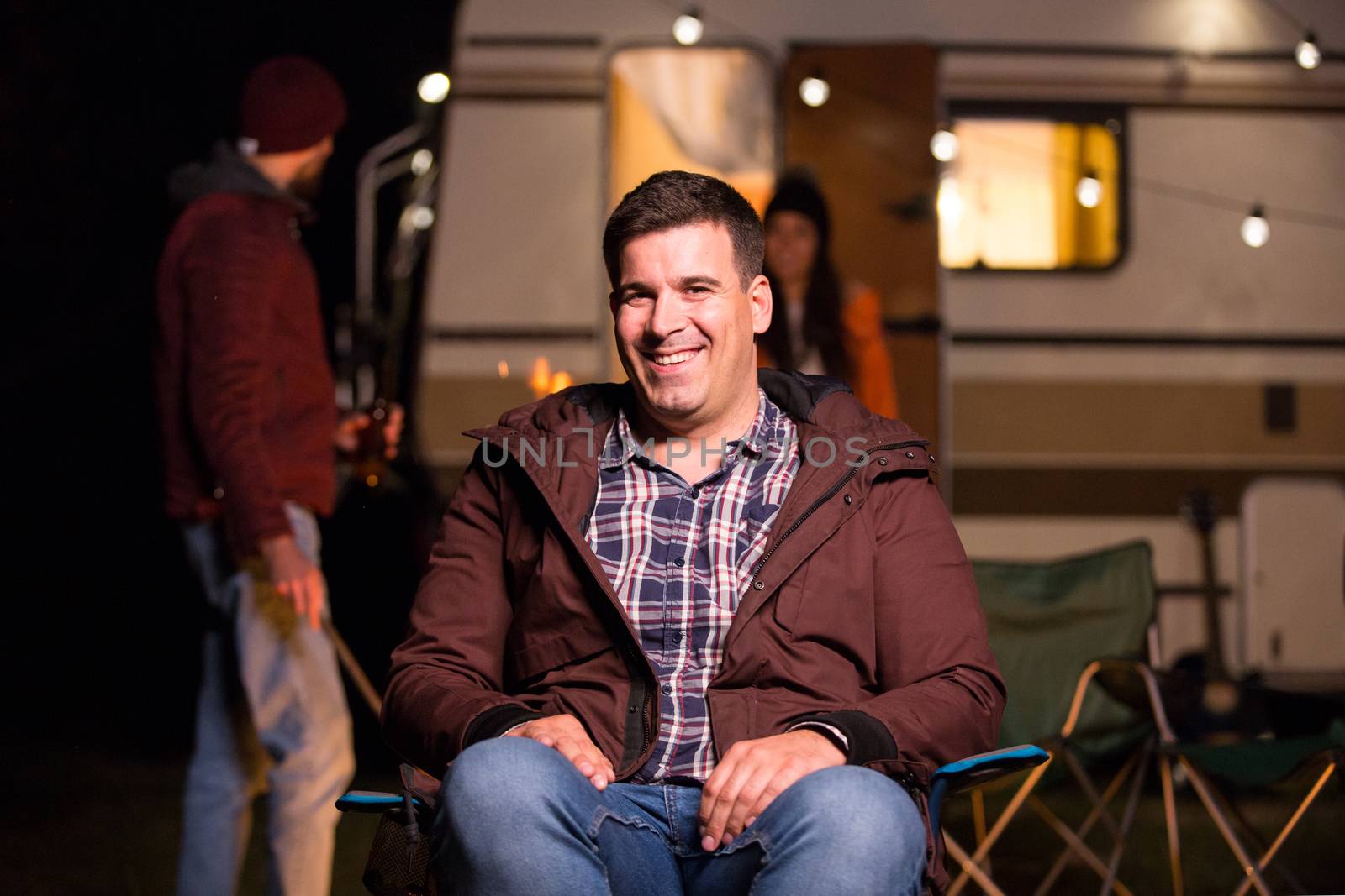 Portrait of tourist sitting on a camping chiar in the mountains with retro camper van in the background. Friends making camp fire stronger.