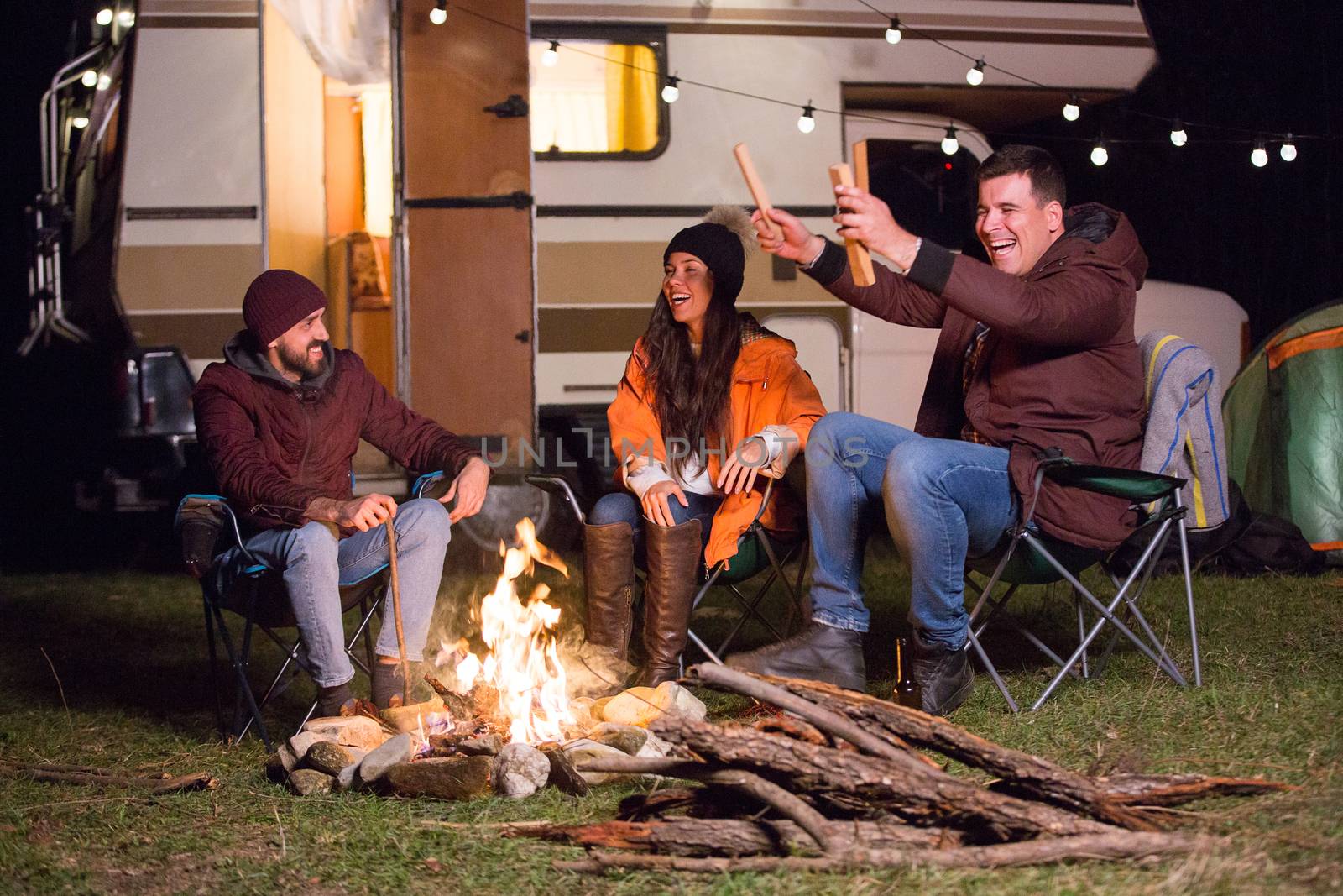 Group of close friends warming up around camp fire and laughing together. Retro camper van.