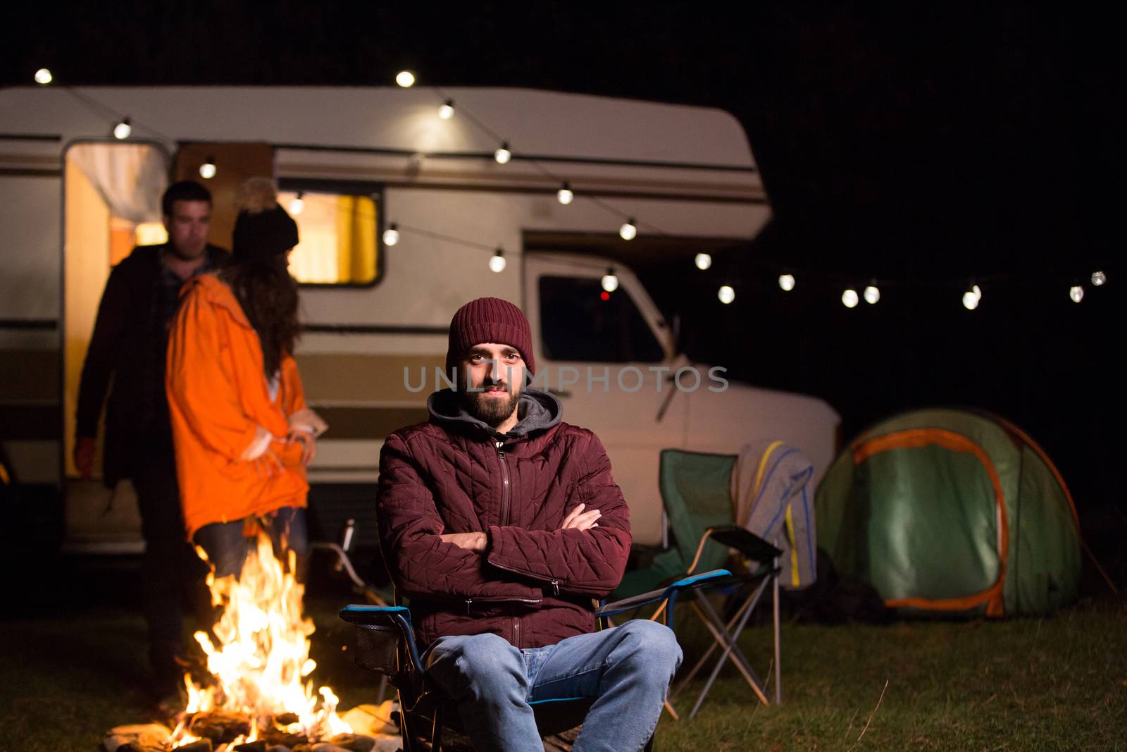 Portrait of bearded tourist with hand crossed in front of camp fire and couple in the background.