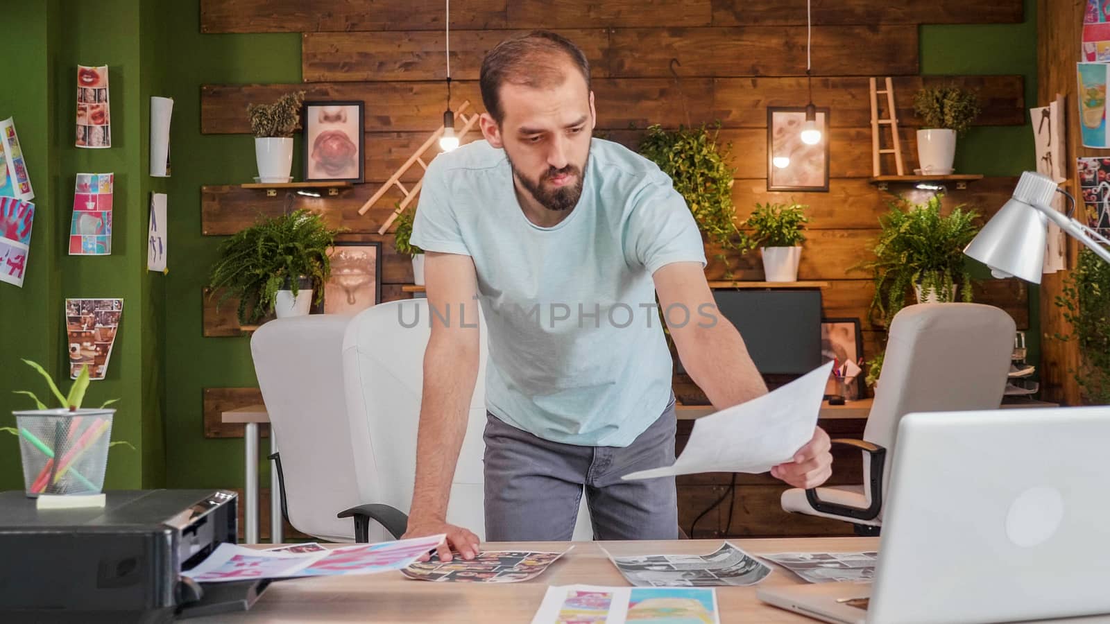 Young designer who is leaning over the table and looks at some pictures printed on the table. Young busy artist