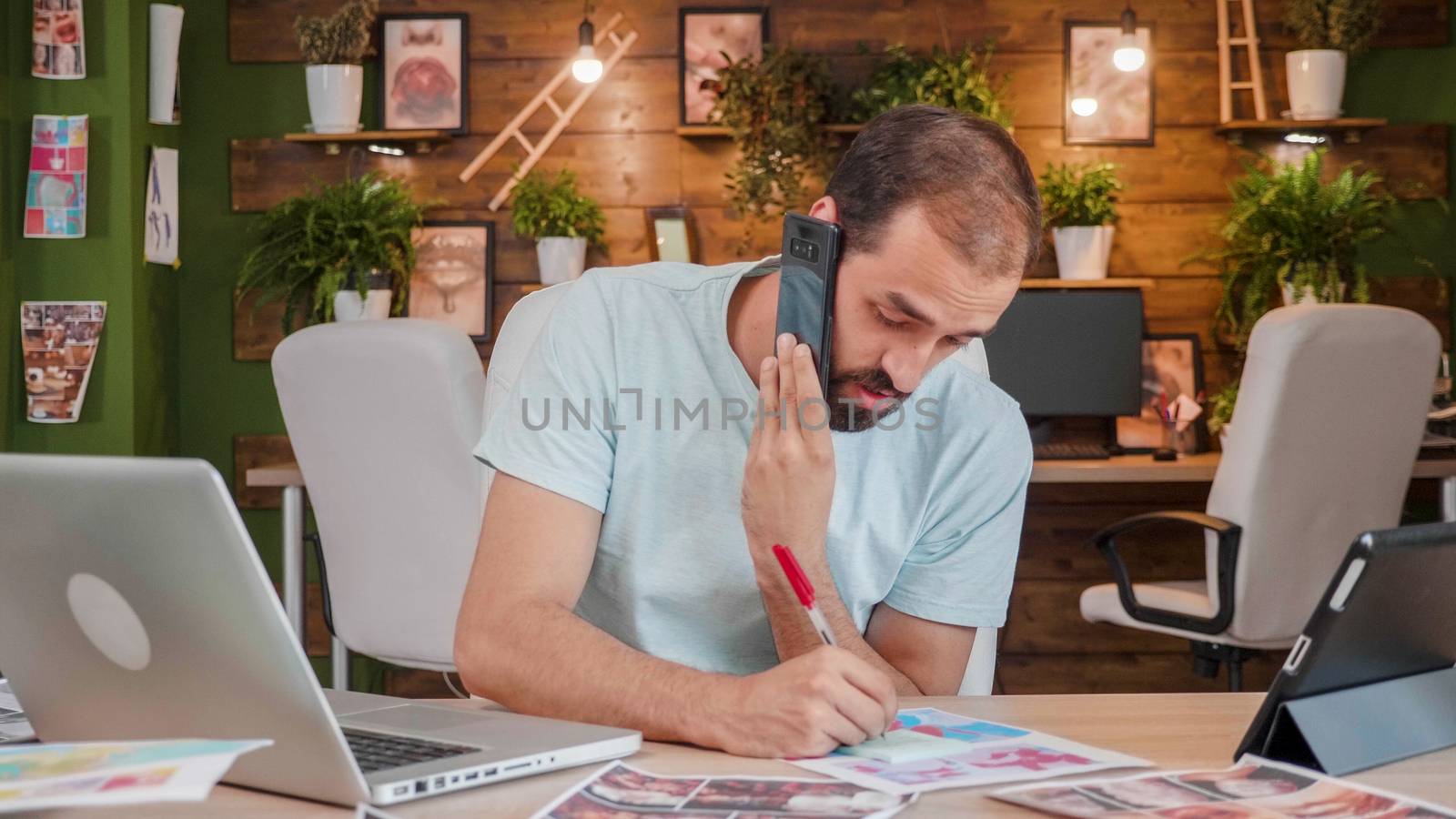 Young designer talking on the cell phone and making some notes on the sheet. Creativ and cozy office