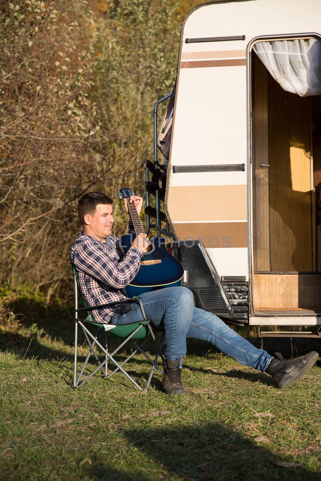 Cheerful young man playing on his guitar in front of his retro camper van in the mountains. Man relaxing in mountains.
