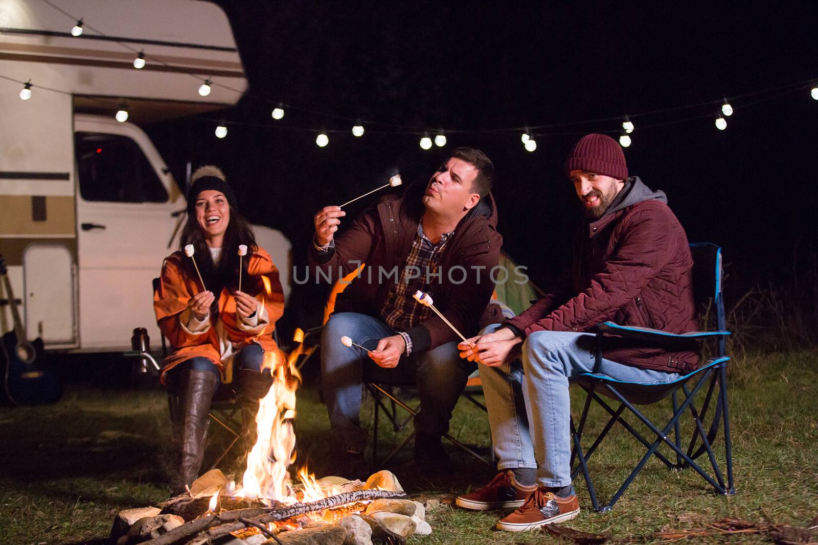 Man blowing a hot marshmallow while friends al laughing around camp fire in the mountains. Retro camper van.