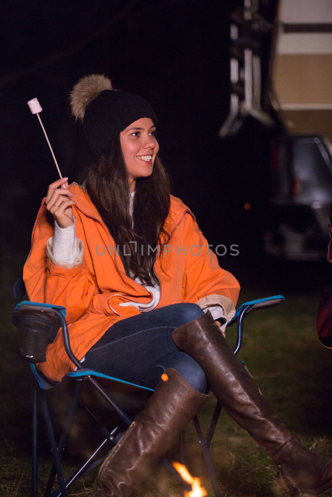 Young girl wearing warm clothes in a cold nigth of autumn in a campsite roasting marshmallow. Retro camper van in the background.