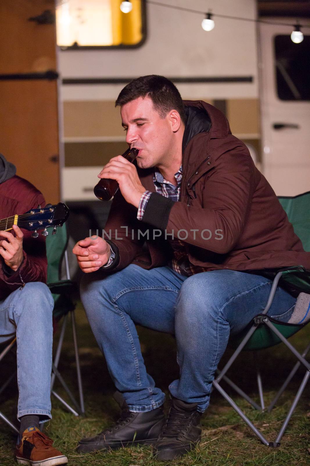 Young man enjoying a bottle of beer in a cold night of autumn in a campsite. Retro camp fire.