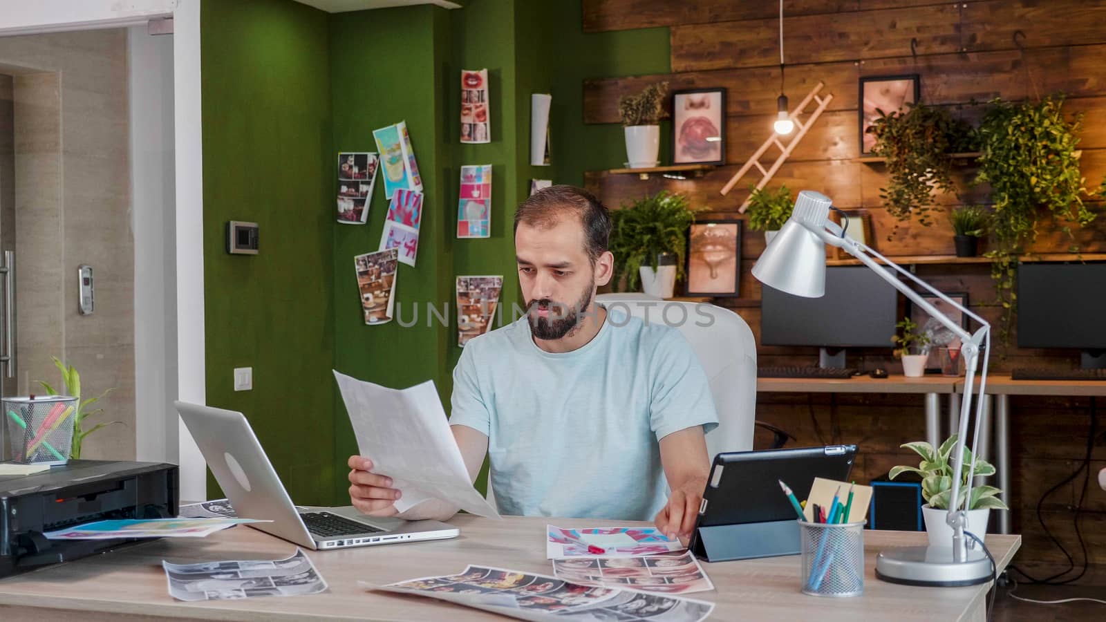 Young designer looking at a sheet holding it in his hand while sitting at the table in front of a laptop. Caucasian creativ artist