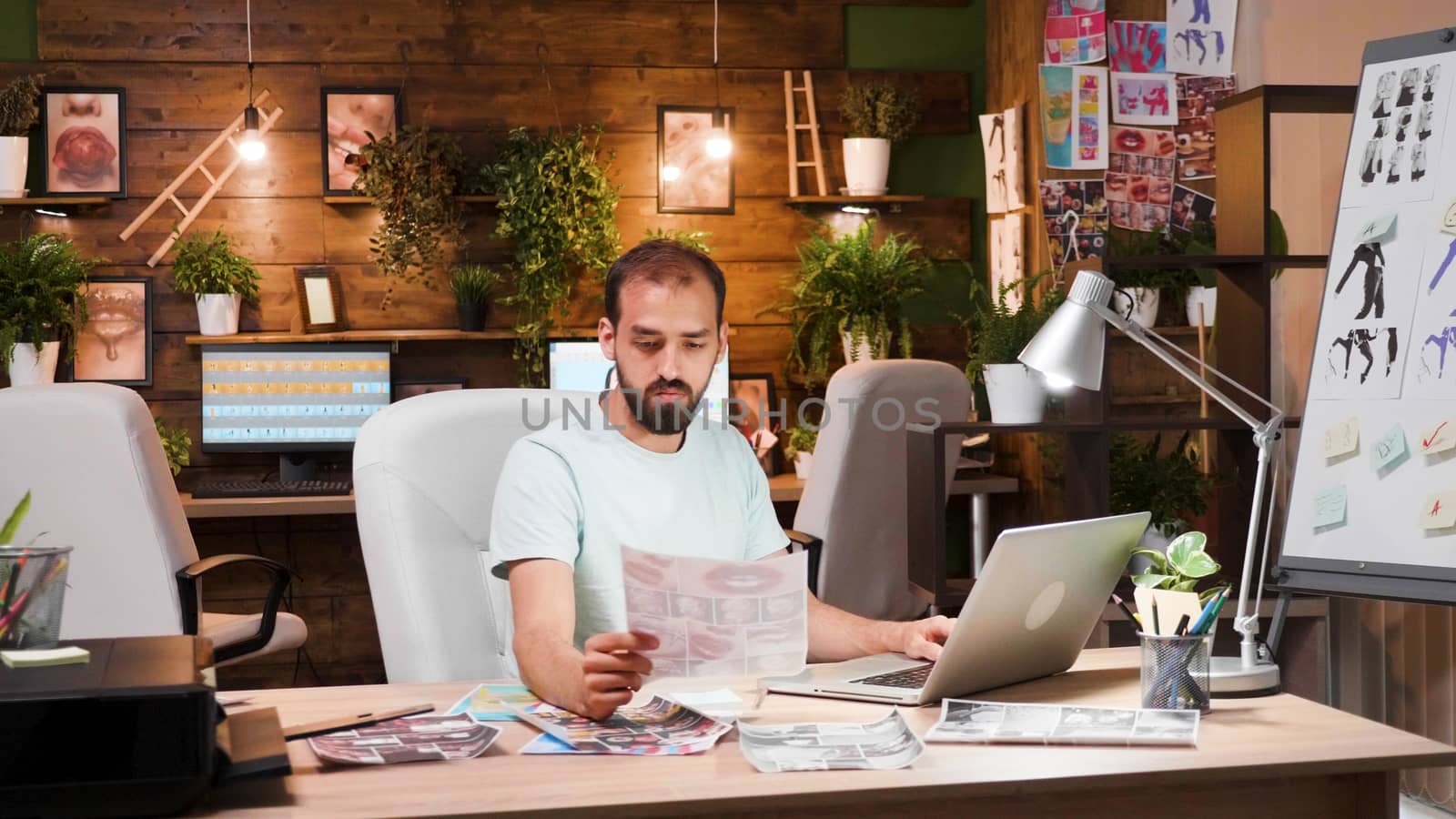 Young designer looking at sheets sitting at his desk with laptop in front. Cozy and smart work space