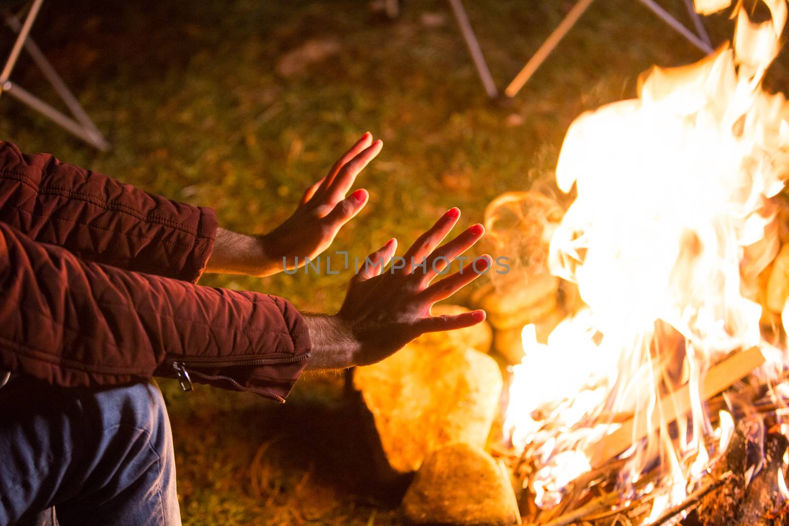 Young man warming up his hands over campfire in a cold night of autumn in the mountains.