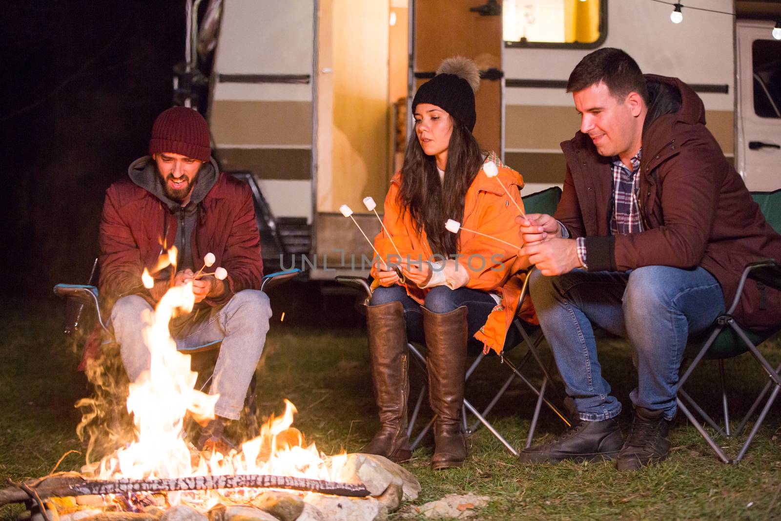 Close friends roasting marshmallows together around camp fire with retro camper van in the background. Mountain campsite.