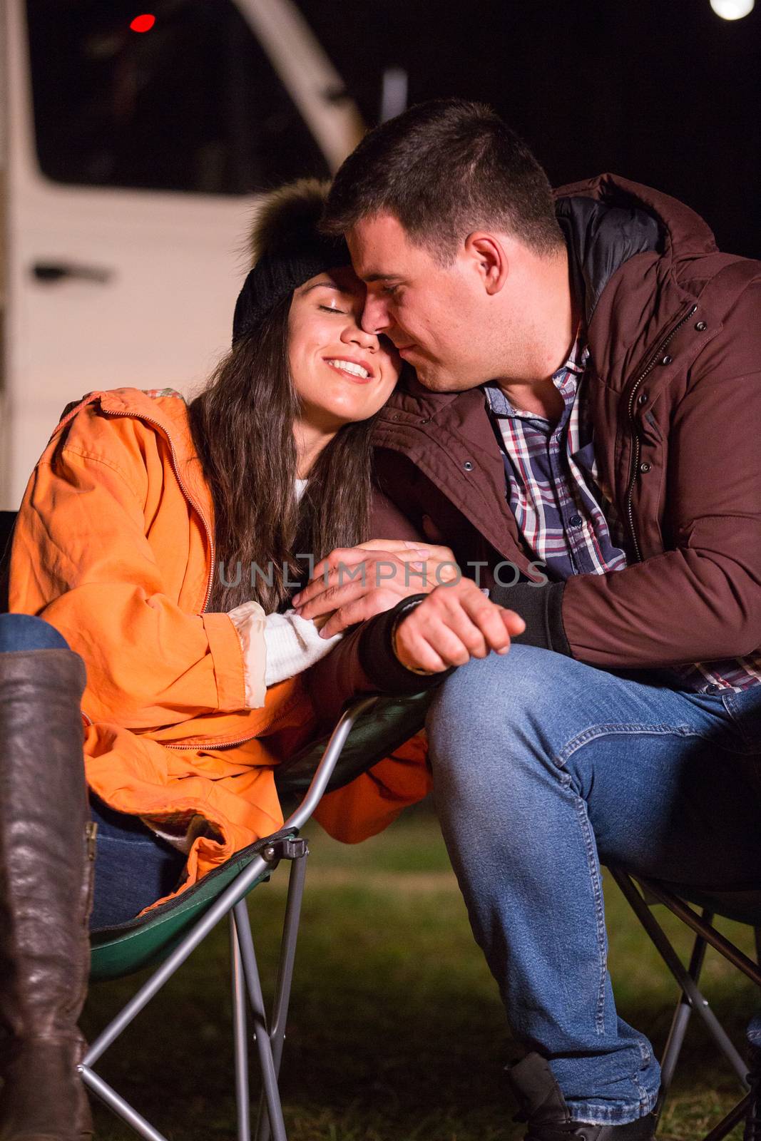 Beautiful couple sharing a sweet moment on the wormth of camp fire in the mountains. Retro camper van in the background.