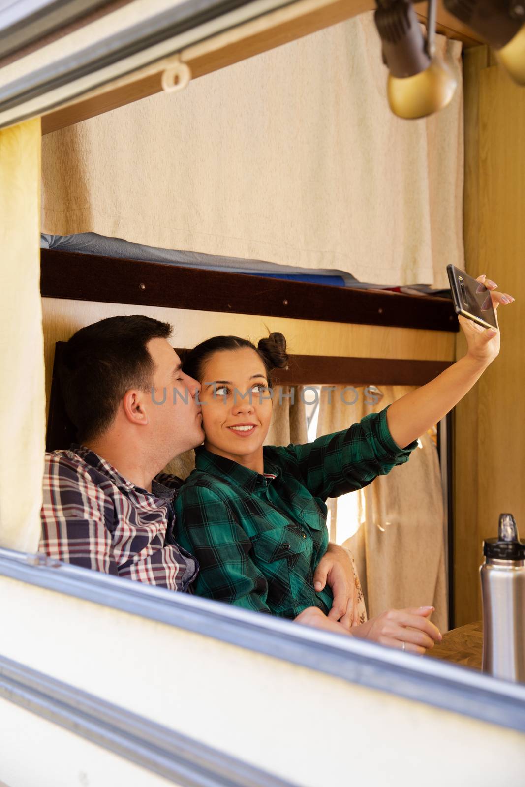 Beautiful young woman taking a selfie inside retro camper van while her boyfriend is kissing her cheeck