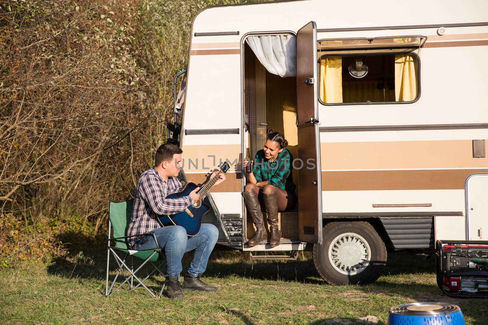 Man trying to play a song on old guitar for his woman in front of their retro camper van. Couple in the mountains.