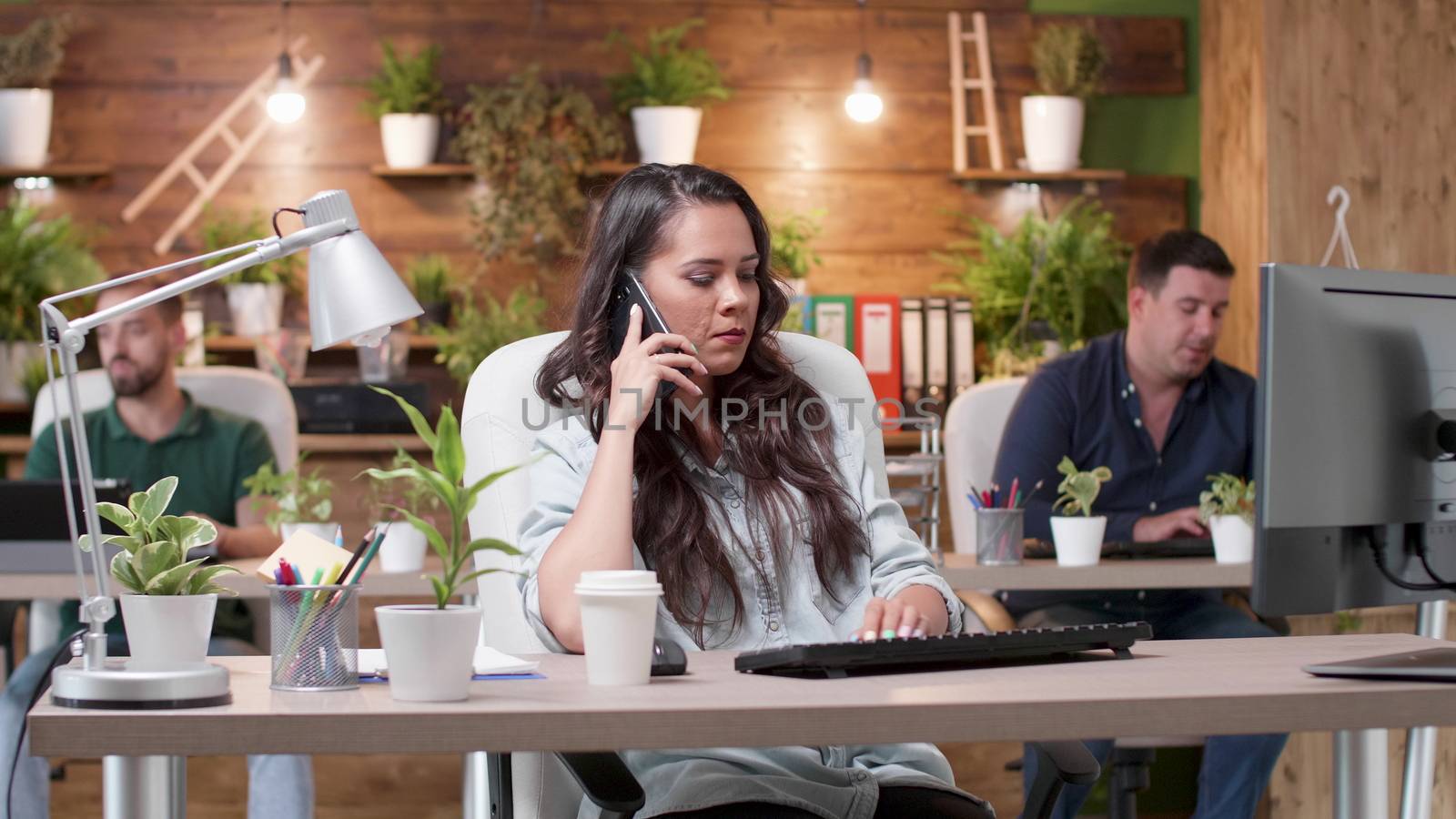 Woman talks on the phone in a busy cozy office. Her colleagues are working in the background