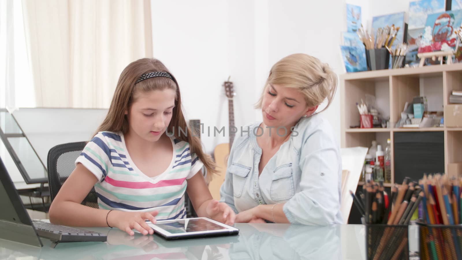 Teenage girl uses a tablet and her mom sits close to her. Mother and daughter looking a tablet screen.