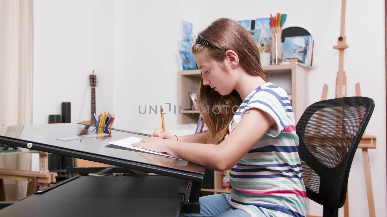 Teenage girl sits at a desk and draws something using a pencil. Teen girl focused on making a drawing.