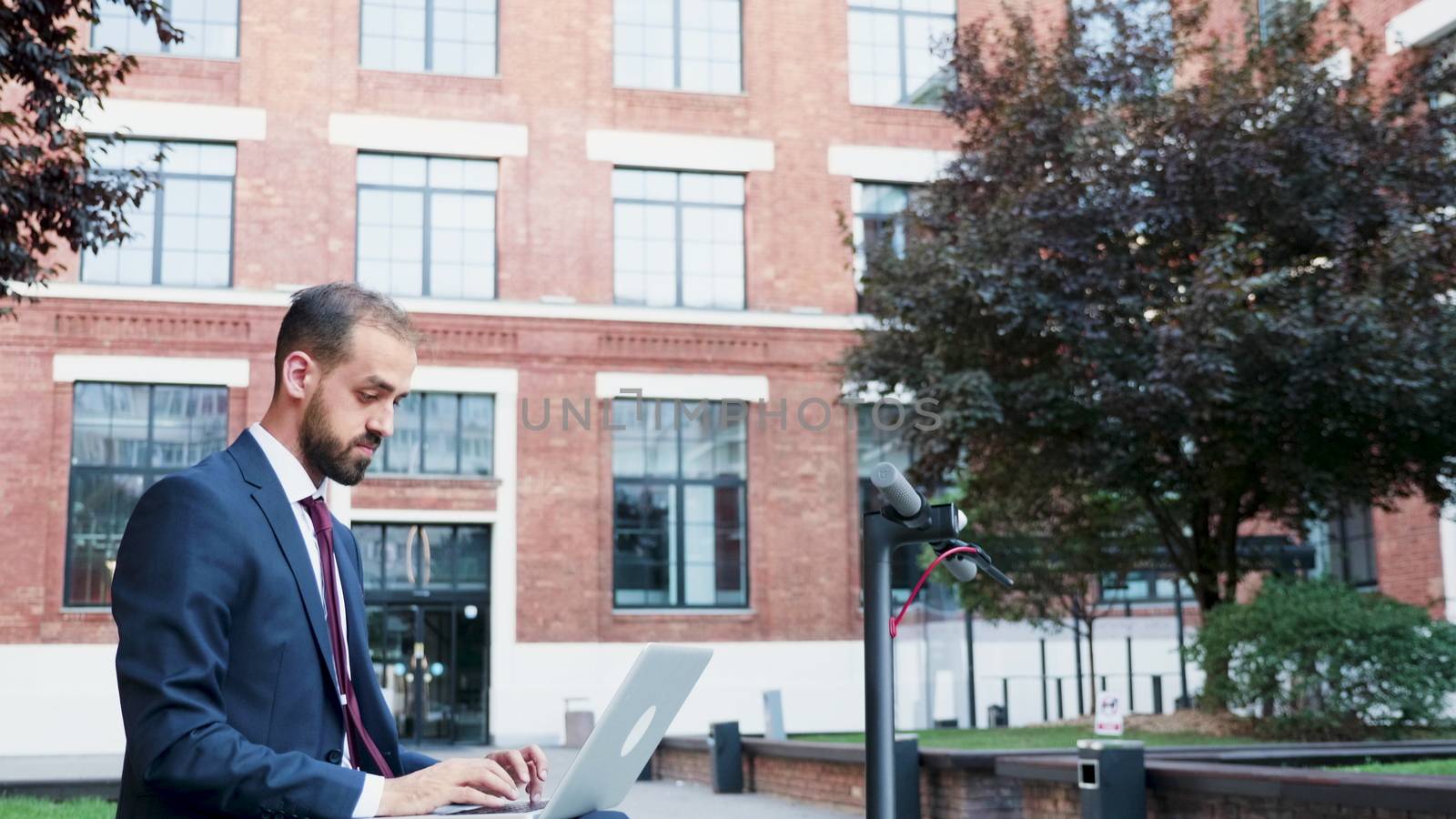 Businessman in formal suit outdoor typing on the laptop sitting in a business district. Panning down shot