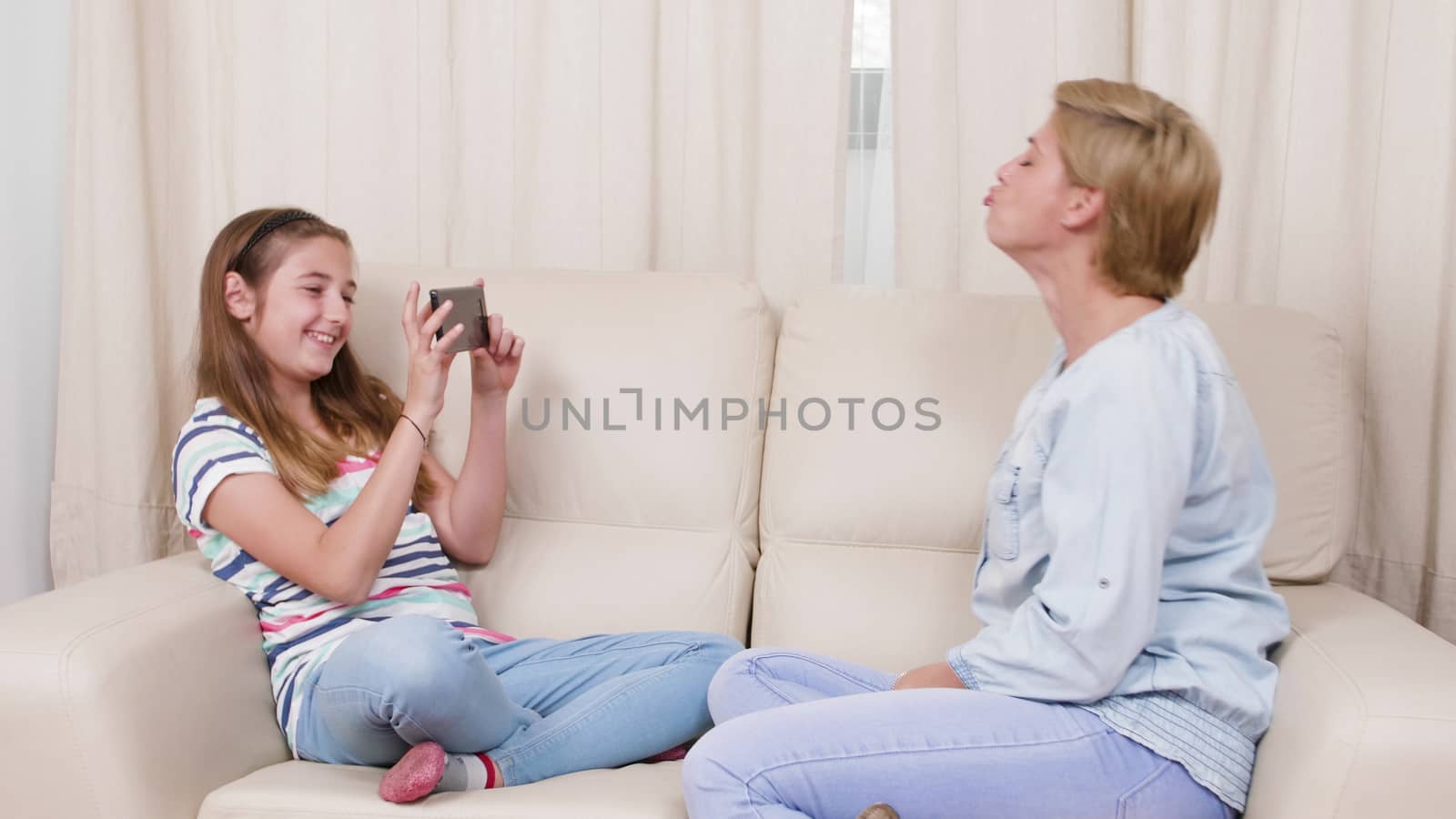 Teenage girl taking pictures of her mother while relaxing on a sofa. Mother and her daughter making photos on a smartphone.