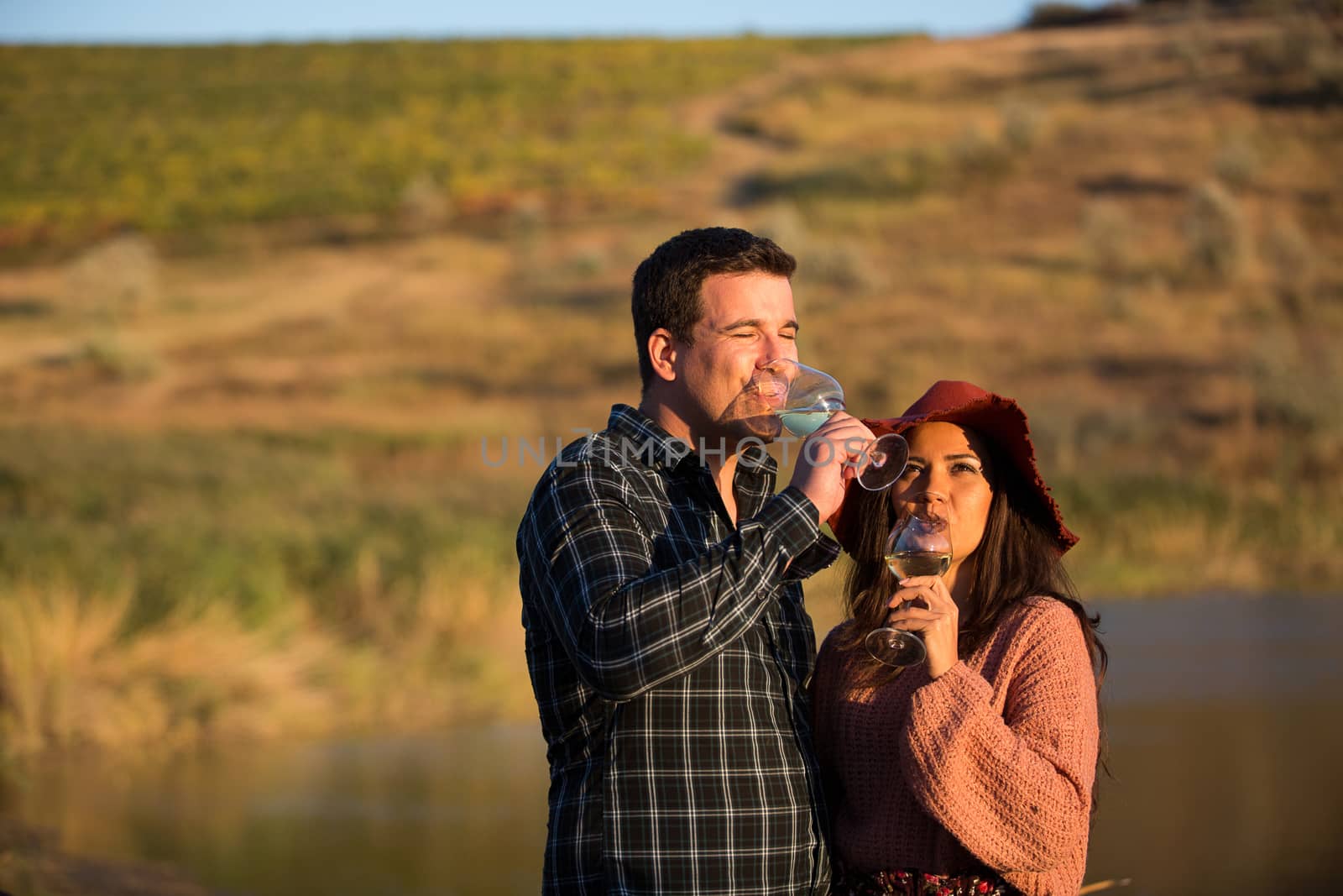 Wine grower couple enjoying a glass of wine in a vineyard with lake behind them at the sunset.