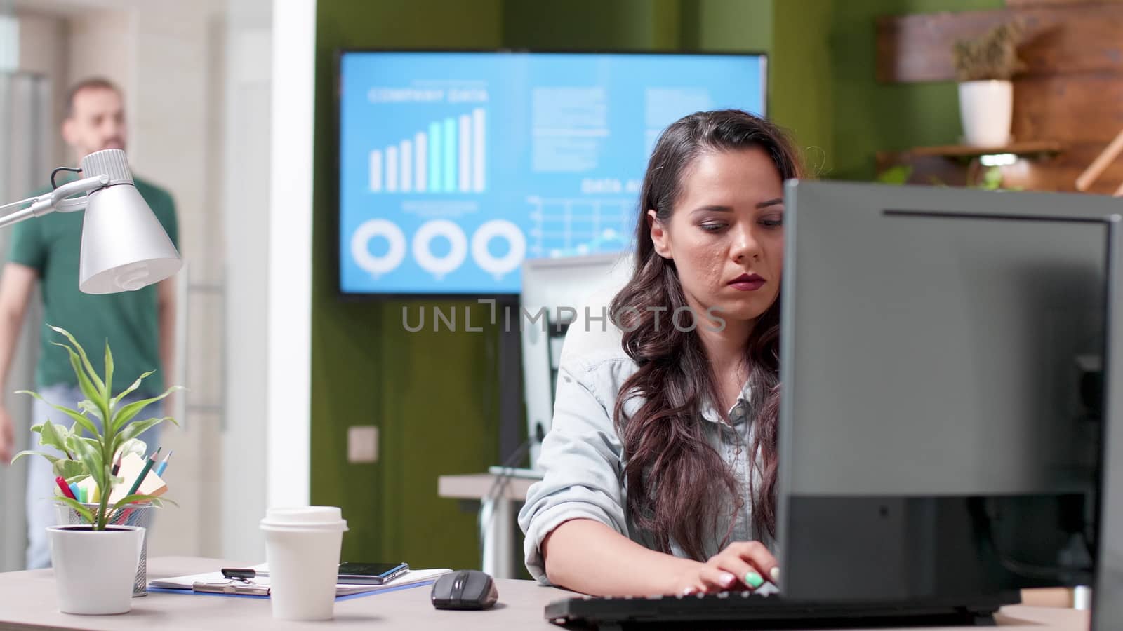 Revealing shot of a busy office with people working in it. A woman in casual clothes sips coffee and types on the computer