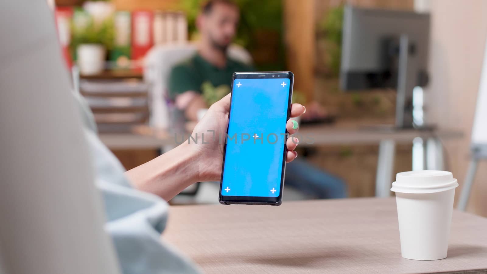 Woman hands holding a smartphone with a blue screen isolated mock up in a busy office