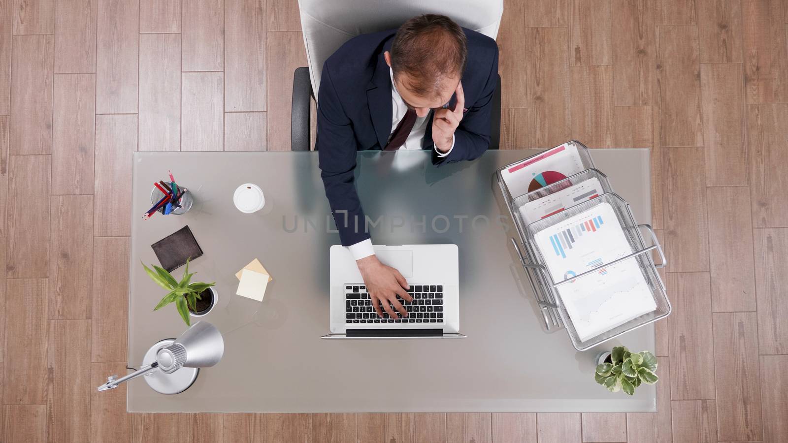 Top view of businessman in suit talking on the phone and typing on laptop sitting at his desk in the office.