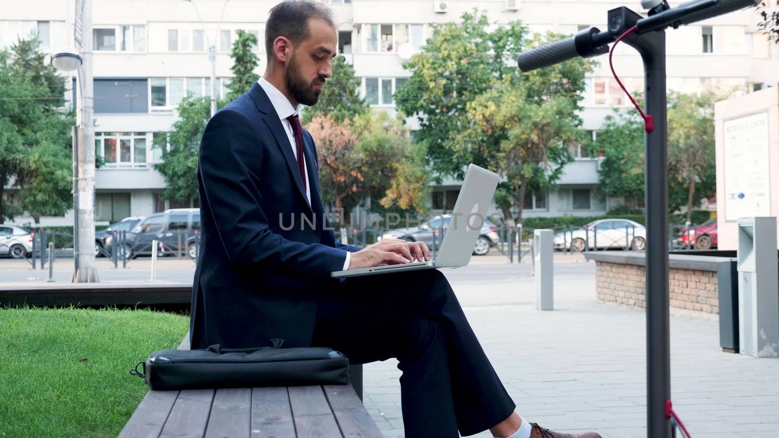 Young manager in suit working on the laptop outdoor in the city, slow motion. Next to him is an electric scooter
