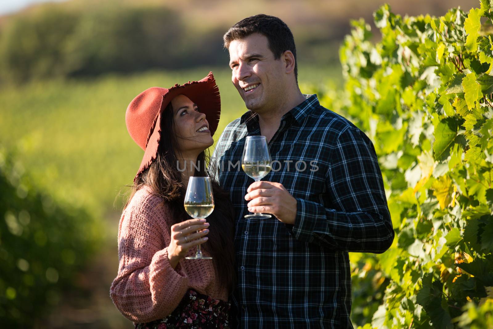 Beautiful young wife holding a glass of wine and looking up at her husband while he is admiring the hills with vine.