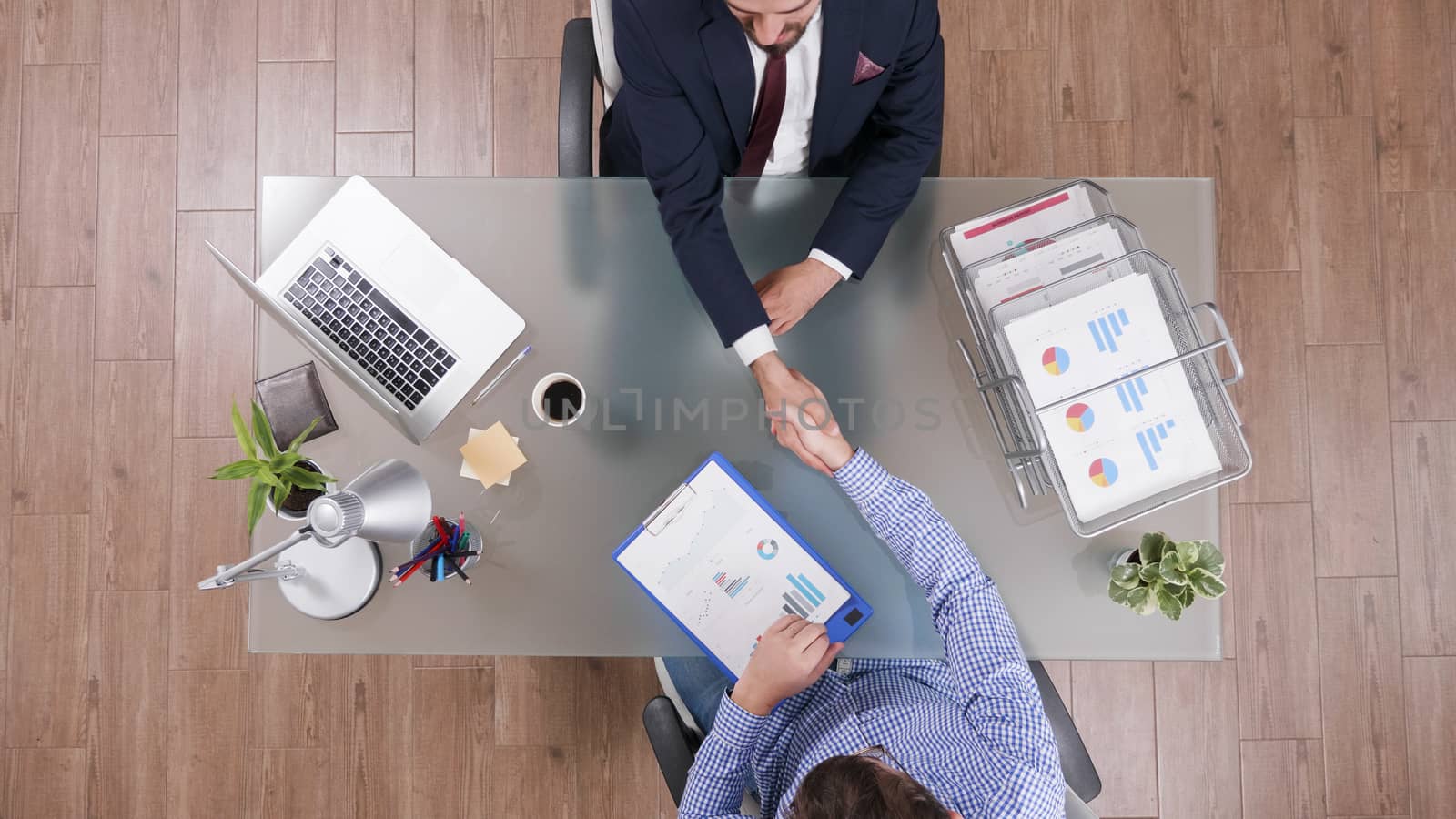 Two businessmen shaking hands sitting at the office with charts on clipboard, top view