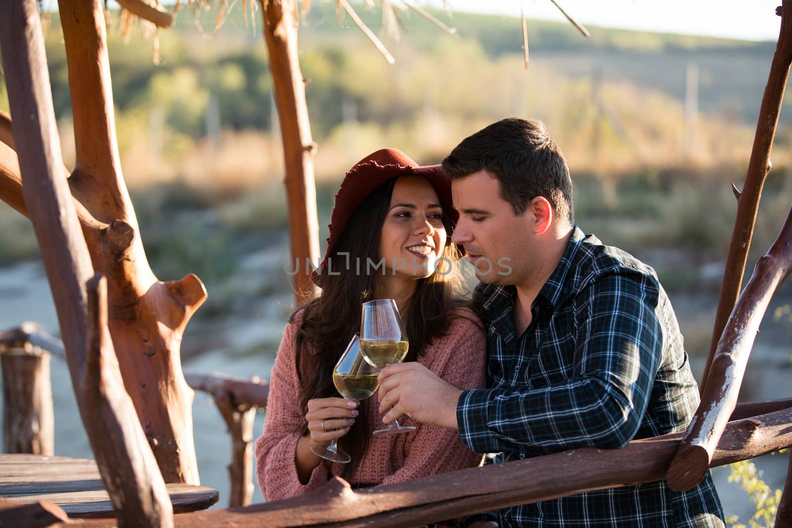 Happy young wife looking at her husband while enjoying together a glass of wine by the edge of a lake in the middle of a vineyard. Beautiful young couple.
