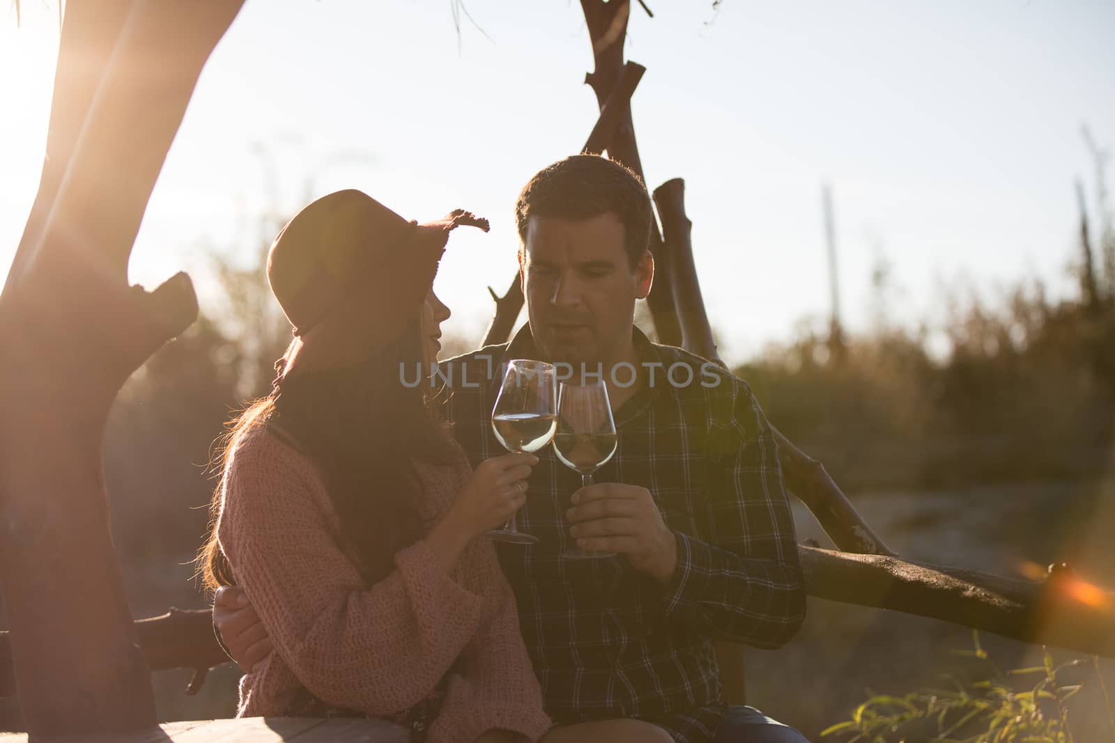 Cheerful young couple having a conversation sitting on a bench in a vineyard clinking a glass of wine.