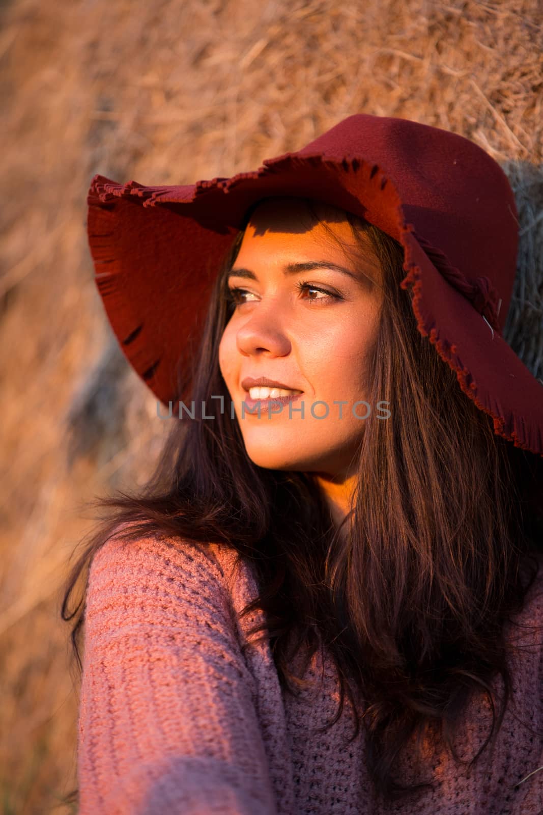 Beautiful young brunette wearing a stylish hat at sunset in a vineyard with straw bale behind her.