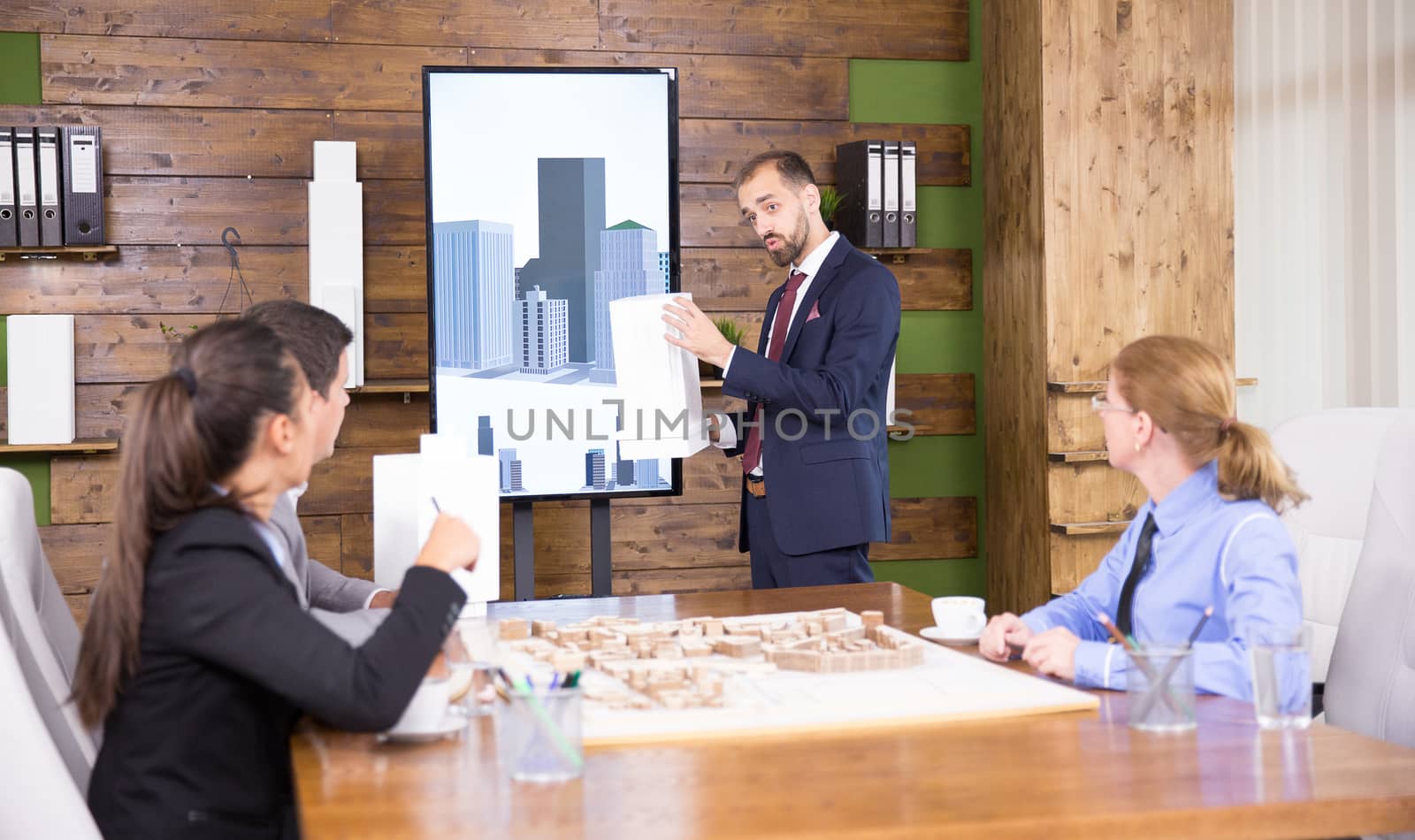 Young architect in business suit holding a building model and presening project to a group of investors.