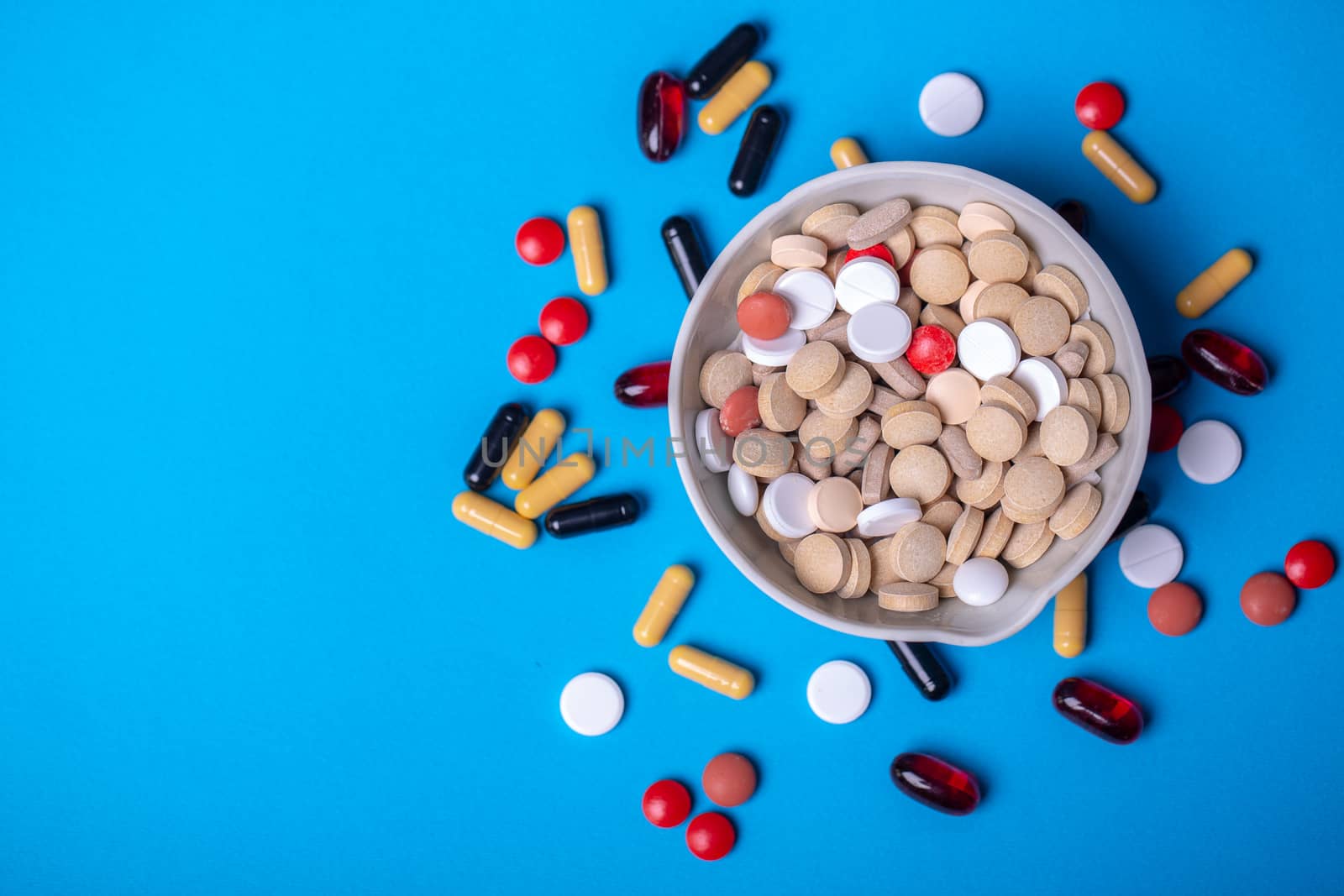 Top view with a bowl of multi-colored pills and next to other pills of different colors and shapes. Copy space and blue background