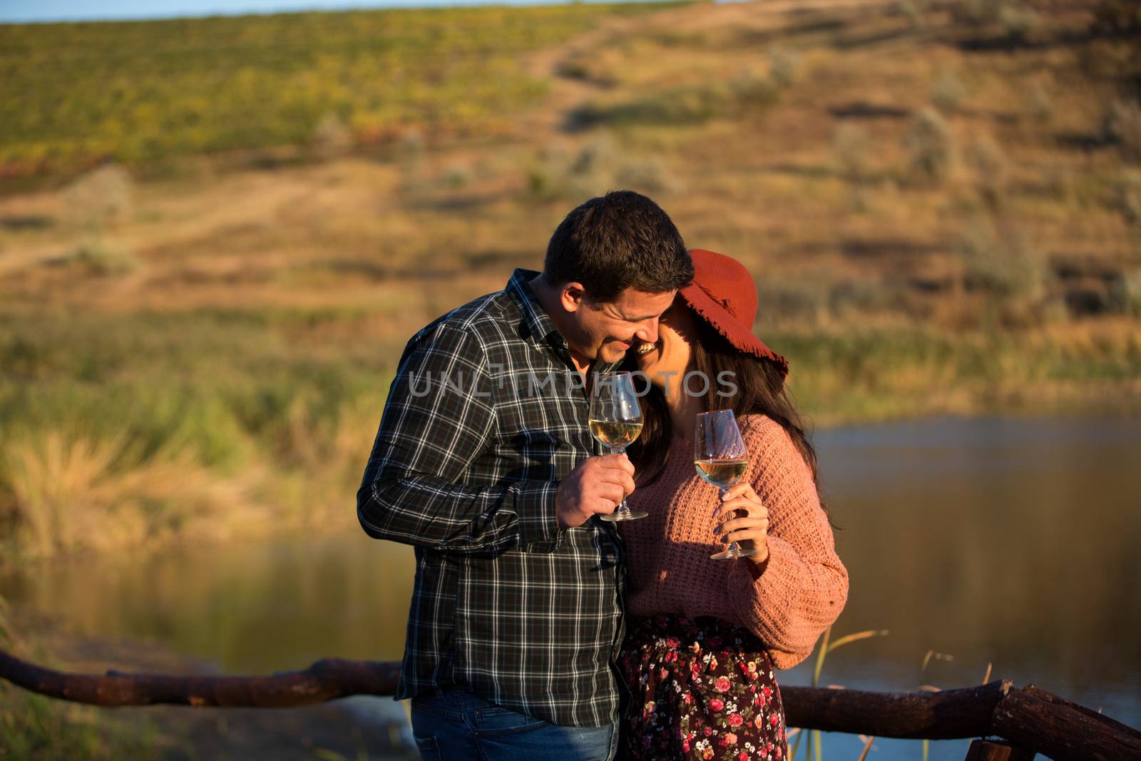 Young wife whispering something funny to her husband ear and holding glasses of wine with lake behind them in the middle of a vineyard.