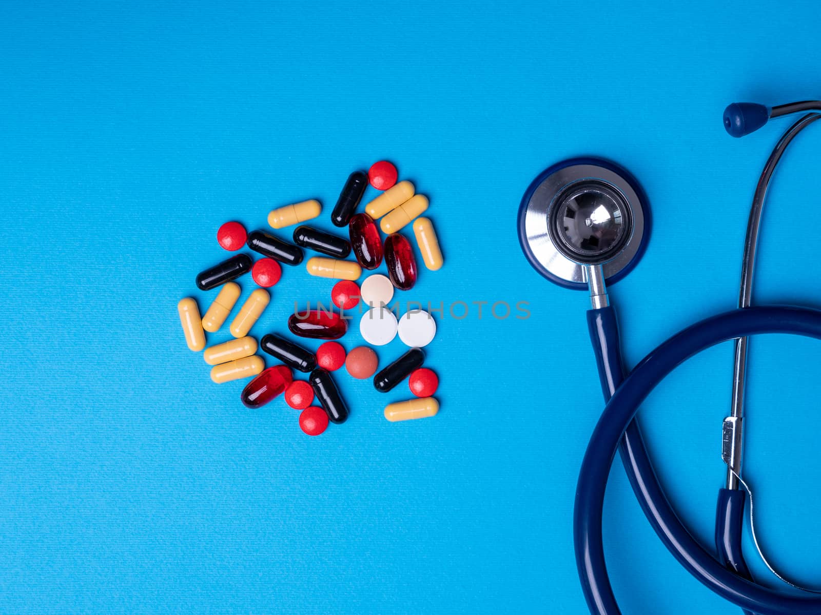 Top view with multicolored pills and capsules next to a stethoscope. Blue background and copy space