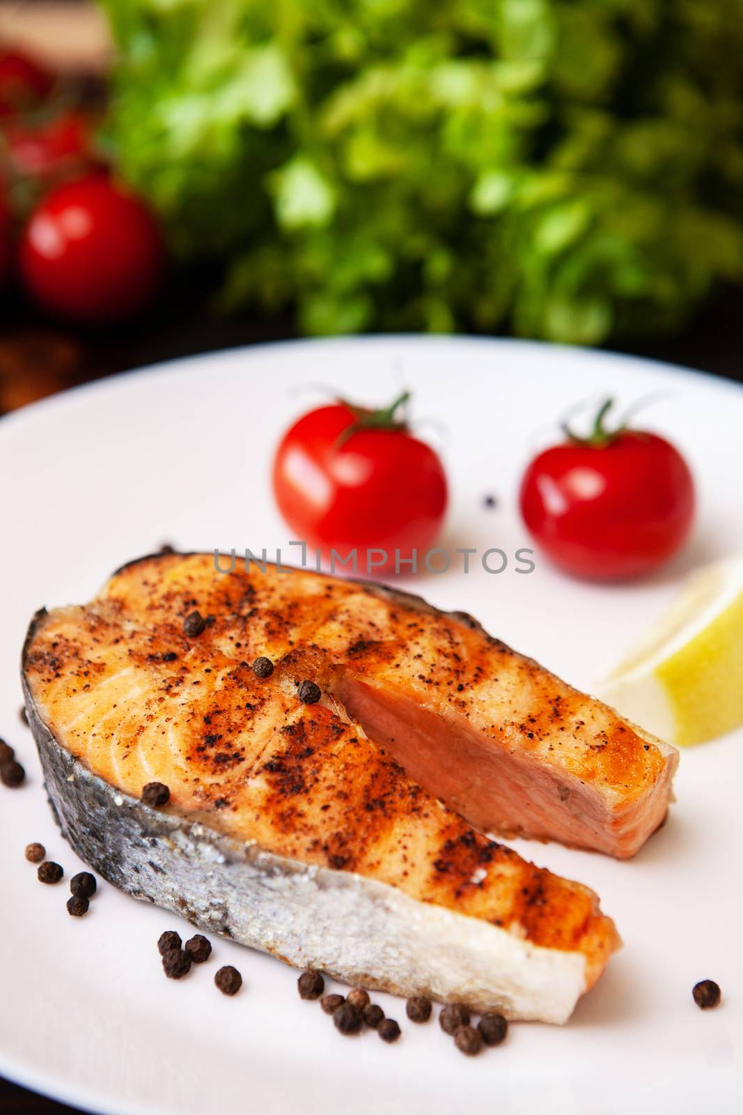 Salmon cooked on the grill arranged on a plate with tomatoes and black pepper. Lemon and salad