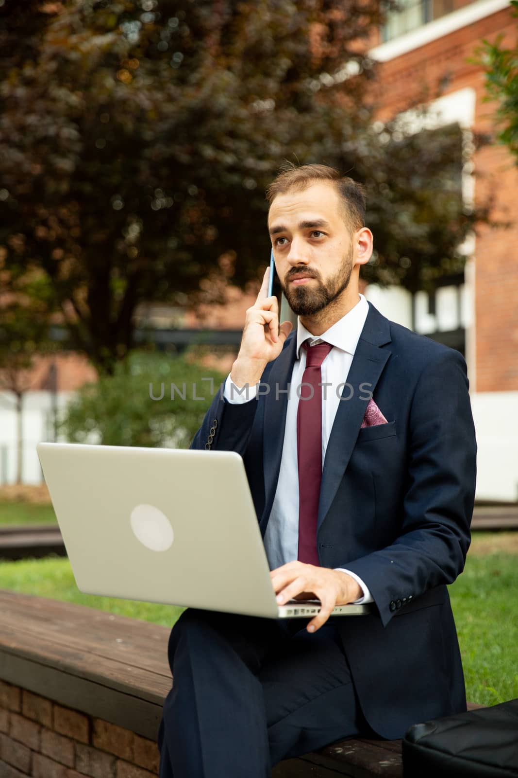 Young businessman working on laptop and talking on the phone in a green space. Suit and business attitude