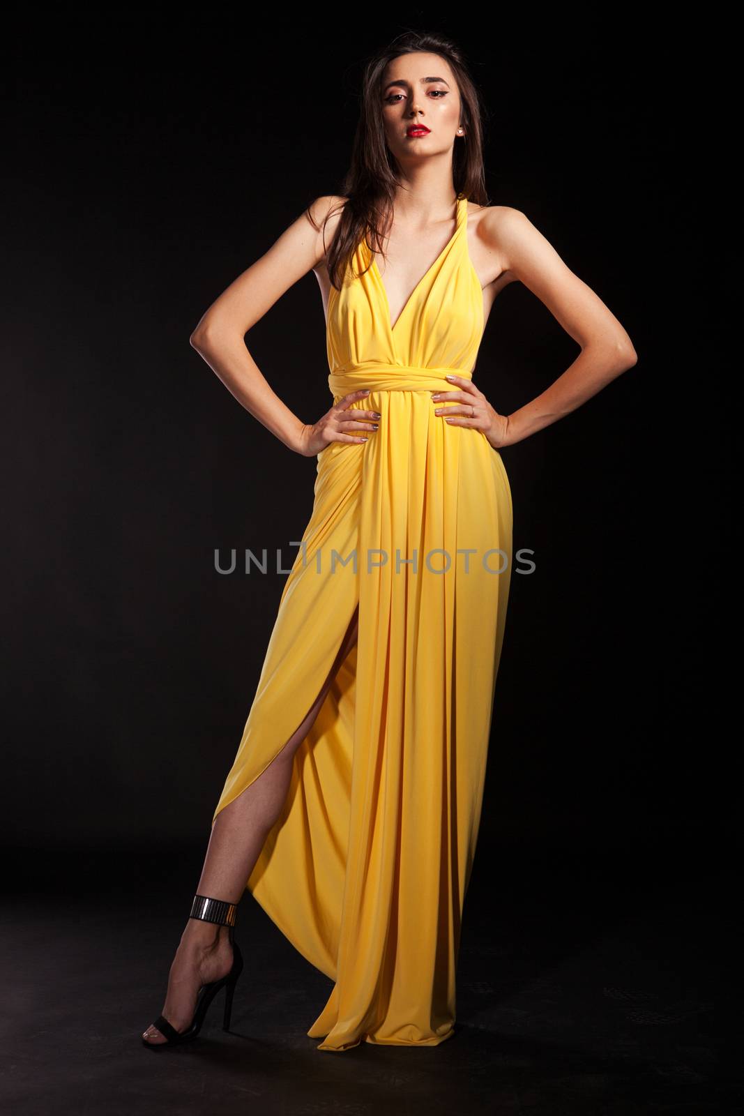 Stylish brunette female wearing yellow fashion dress on black background holding hands on her hips.