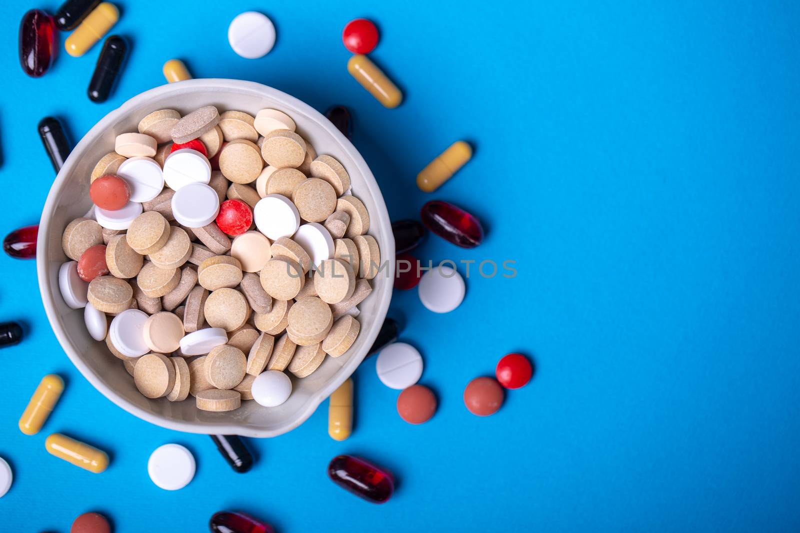 Top view with a red pill in a bowl with pills and next to other pills of different colors and shapes. Copy space and blue background