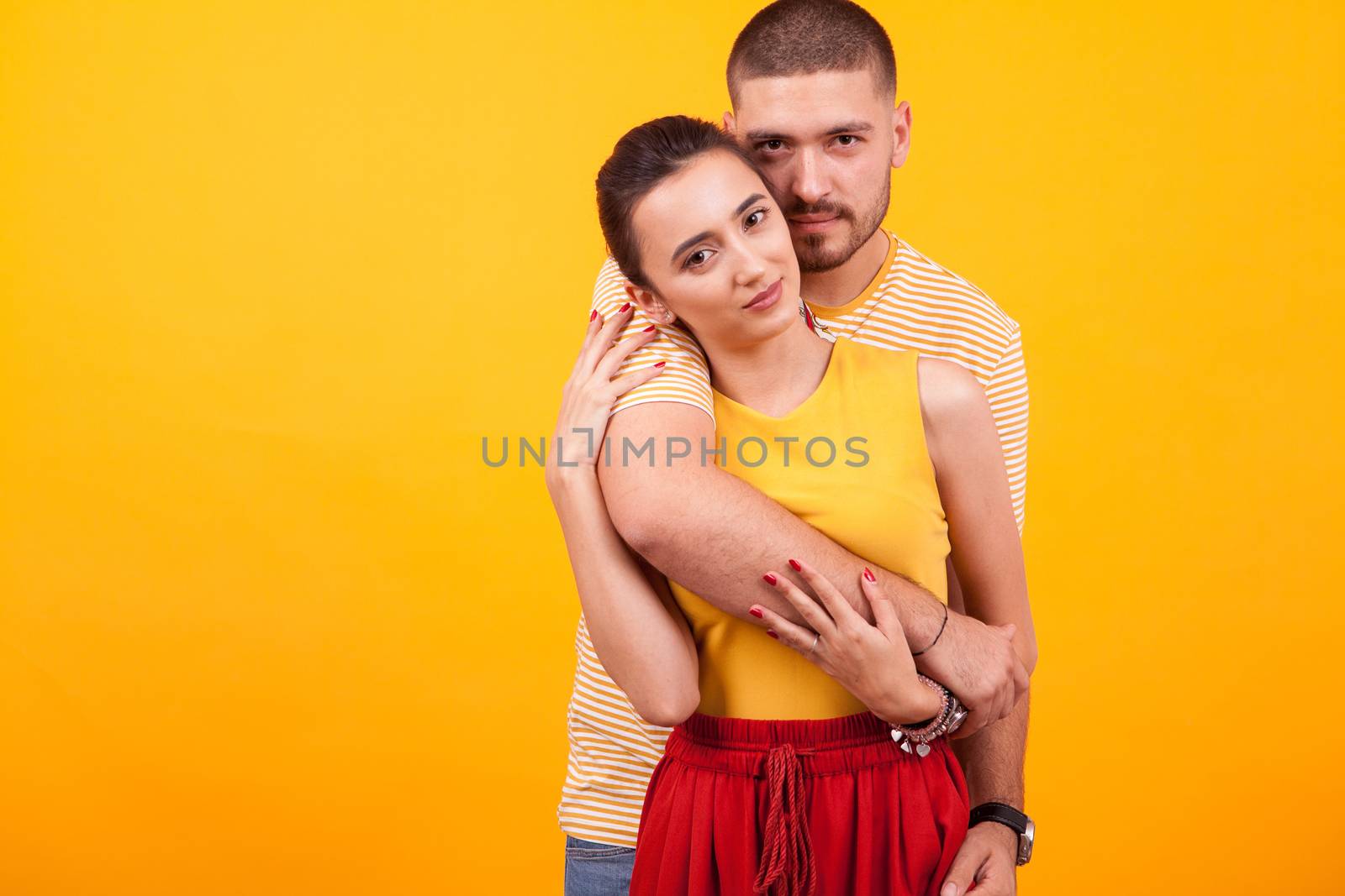 Handsome boyfriend hugging his beautiful girlfriend from behind in studio over yellow background.