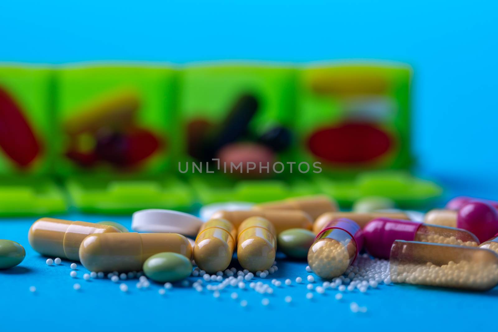 Top view with multicolored pills and capsules and in the back you see an organizer. Green organizer and blue background